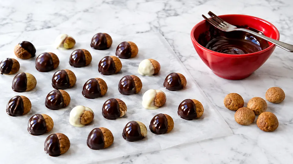 Pebernødder dyppet halvt i mørk og hvid chokolade på bagepapir ved siden af en rød skål med smeltet chokolade
